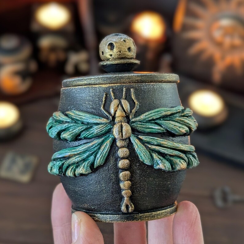 May include: A small black lidded jar featuring a textured surface with a prominent three-dimensional dragonfly relief. The dragonfly has metallic green wings and a gold-toned body. A spherical knob with a cratered texture sits on the lid.
