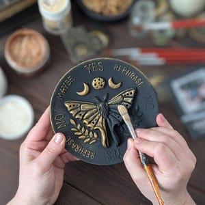 May include: A circular black plaque featuring a raised gold moth at the center, surrounded by celestial symbols including a moon and stars. The text YES, NO, MAYBE, and REPHRASE is embossed around the perimeter in gold lettering.