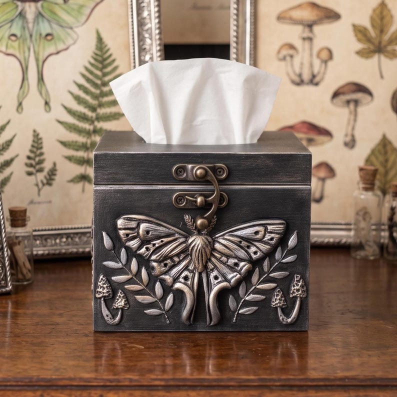 Puede incluir: Una caja de madera gris oscuro para pa&ntilde;uelos con un emblema de polilla en relieve de color plateado en el panel frontal. La caja incluye un cierre de metal y tallas decorativas plateadas de ramas con hojas y peque&ntilde;os hongos.