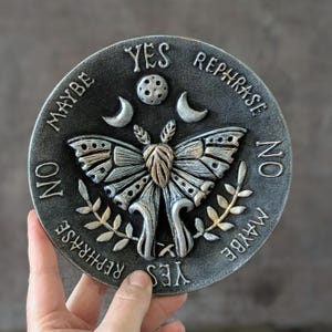 May include: A circular gray plaque featuring a central moth relief with raised wings, flanked by two crescent moons and a small circle. The text YES, NO, MAYBE, and REPHRASE is embossed around the perimeter in a decorative font style.
