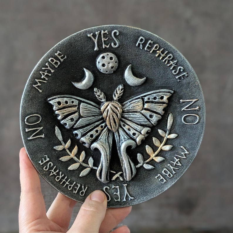 May include: A circular gray plaque featuring a central moth relief with raised wings, flanked by two crescent moons and a small circle. The text YES, NO, MAYBE, and REPHRASE is embossed around the perimeter in a decorative font style.