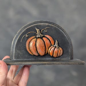 May include: A semi-circular black decorative plaque featuring a raised relief design of two orange pumpkins with textured stems and curling vines. The piece has a distressed finish with visible gold-toned highlights along the edges and details.