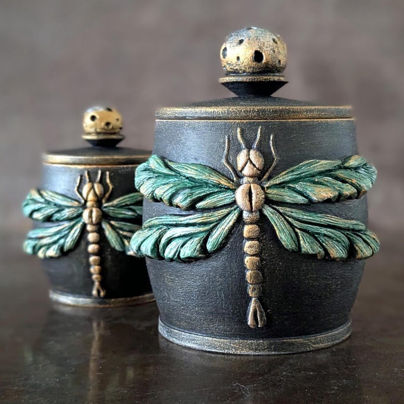 May include: Two cylindrical black jars with lids, each featuring a raised, three-dimensional dragonfly motif. The dragonflies have textured green wings and gold-toned bodies. Each lid is topped with a small, round, gold-speckled knob.