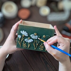 May include: A green wooden box with white and gold floral relief details. The box is being painted with a small brush. The flowers have white petals and gold stems and leaves. The box is held by two hands.
