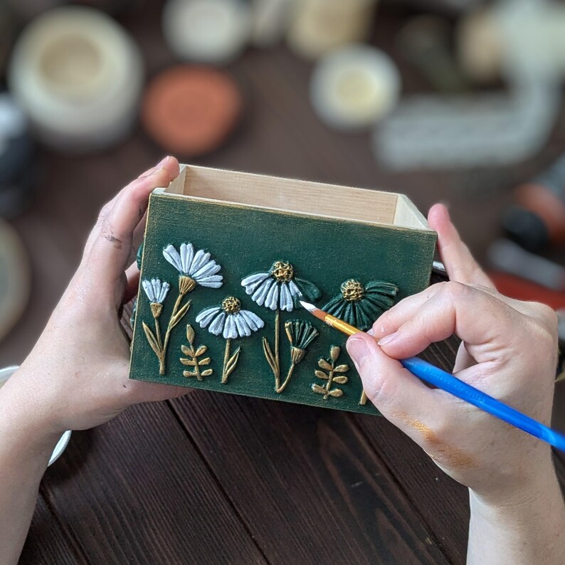 May include: A green wooden box with white and gold floral relief details. The box is being painted with a small brush. The flowers have white petals and gold stems and leaves. The box is held by two hands.