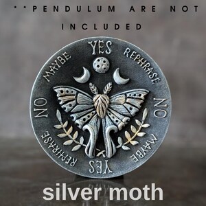 May include: A circular silver-toned metal divination board featuring a central moth with raised wings, flanked by crescent moons and a moon phase. The text reads YES, NO, MAYBE, and REPHRASE. The top text states PENDULUM ARE NOT INCLUDED.