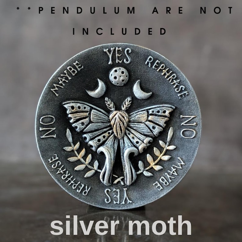 May include: A circular silver-toned metal divination board featuring a central moth with raised wings, flanked by crescent moons and a moon phase. The text reads YES, NO, MAYBE, and REPHRASE. The top text states PENDULUM ARE NOT INCLUDED.