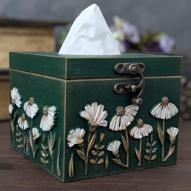 May include: A dark green wooden tissue box with a hinged lid and metal clasp. The box is decorated with raised white and gold floral designs. White tissues are visible at the top.