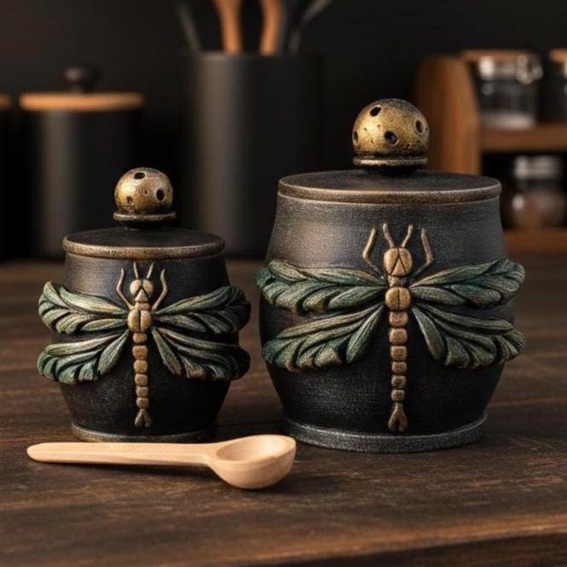May include: Two dark charcoal-colored ceramic canisters with lids, each featuring a raised, textured dragonfly motif with muted green wings and a bronze-toned body. A small, light-colored wooden spoon rests horizontally in front of the canisters.