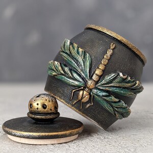 May include: A black cylindrical container with a textured surface featuring a raised, sculpted dragonfly with metallic green wings and a gold body. The container includes a matching black lid with a gold-accented, perforated knob handle.