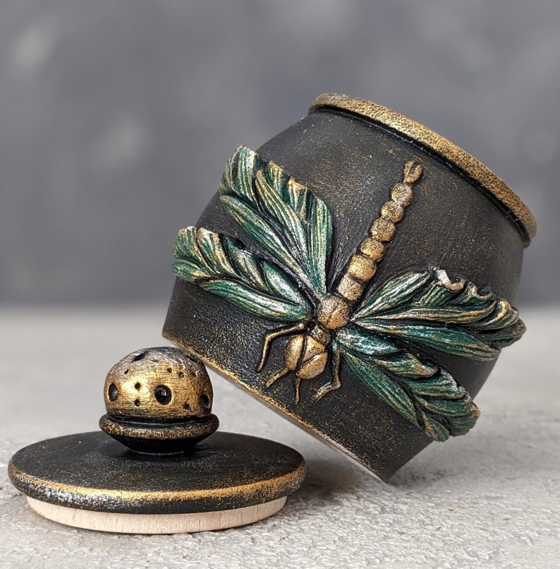 May include: A black cylindrical container with a textured surface featuring a raised, sculpted dragonfly with metallic green wings and a gold body. The container includes a matching black lid with a gold-accented, perforated knob handle.