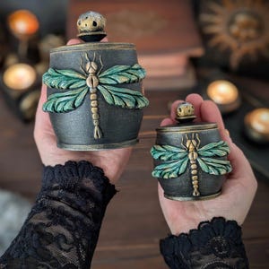 May include: Two cylindrical containers with lids, featuring a textured black finish and a prominent relief of a dragonfly with metallic green wings and a gold body. Each lid is topped with a small, round, speckled knob in gold and black.