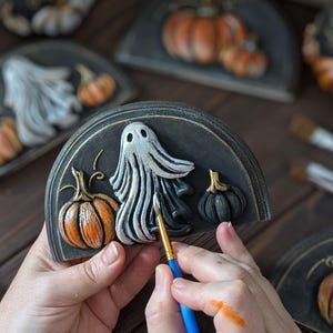 May include: A semi-circular black plaque featuring a raised white ghost figure in the centre, flanked by a textured orange pumpkin on the left and a smaller black pumpkin on the right. A blue-handled paintbrush is applying white paint to the ghost.