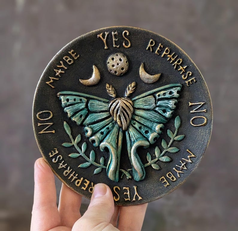 May include: A circular dark bronze plaque featuring a central teal and gold moth with raised wings. Above the moth are a moon and stars. The text YES, NO, MAYBE, and REPHRASE is embossed around the perimeter in a gold finish on the dark surface.