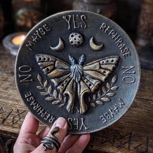 May include: A circular bronze-toned plaque featuring a central moth relief, flanked by crescent moons and a small circle. The text YES, NO, MAYBE, and REPHRASE is embossed around the perimeter. A snake-shaped ring is worn on a finger.