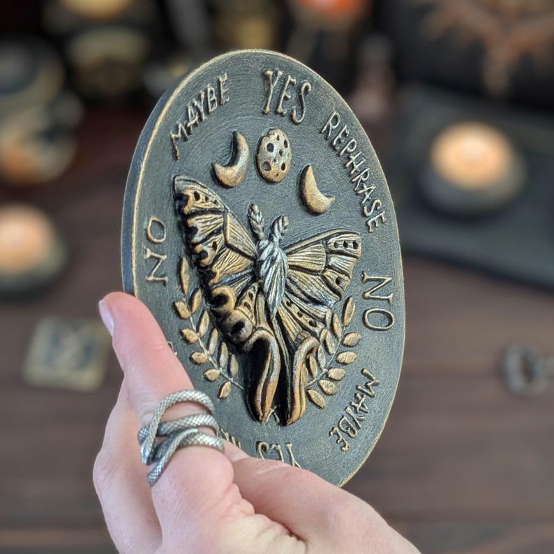 May include: An oval black and gold plaque featuring a central moth relief, moon phases, and leaf motifs. The text reads YES, NO, MAYBE, and REPHRASE arranged around the perimeter. The surface has a weathered, metallic finish with raised lettering.
