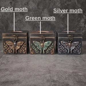 Puede incluir: Tres cajas de pa&ntilde;uelos cuadradas de madera con dise&ntilde;os de polillas en relieve en el frente. La caja izquierda tiene una polilla dorada, la central una verde y la derecha una plateada. Cada caja incluye motivos de hojas y setas.