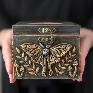 Puede incluir: Una caja rectangular de madera con un acabado oscuro y desgastado que presenta un relieve de polilla en tono dorado en el centro. El dise&ntilde;o incluye motivos en relieve de setas y hojas en el panel frontal, asegurada con un cierre met&aacute;lico.