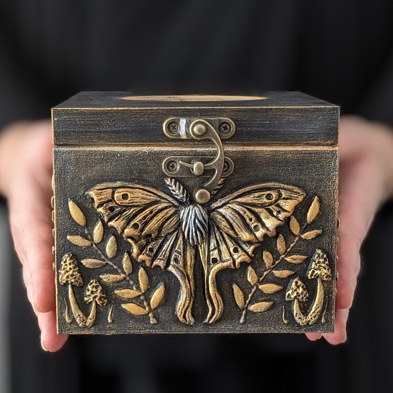Puede incluir: Una caja rectangular de madera con un acabado oscuro y desgastado que presenta un relieve de polilla en tono dorado en el centro. El dise&ntilde;o incluye motivos en relieve de setas y hojas en el panel frontal, asegurada con un cierre met&aacute;lico.