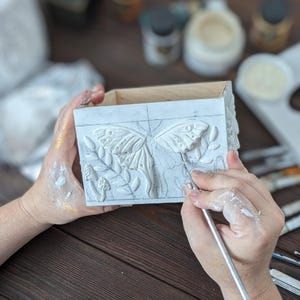 Puede incluir: Una caja rectangular de madera con un relieve blanco de una polilla y setas en el panel frontal. Se utiliza un pincel fino para aplicar pintura blanca sobre la superficie texturizada, que tambi&eacute;n muestra tenues bocetos a l&aacute;piz.