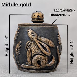 Puede incluir: Un tarro decorativo negro con un dise&ntilde;o de conejo dorado y tapa. El tarro mide aproximadamente 10 cm de alto y 6,6 cm de di&aacute;metro. El tarro tiene una altura de 8,1 cm. La tapa tiene un dise&ntilde;o perforado. El texto "Middle gold" es visible.