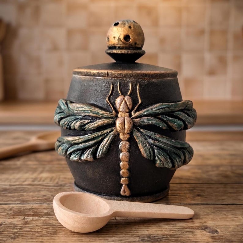 May include: A black ceramic jar with a lid featuring a textured gold-toned knob. The jar is decorated with a raised, sculpted dragonfly motif in shades of dark green and gold. A small, light-colored wooden spoon is placed in front of the jar.