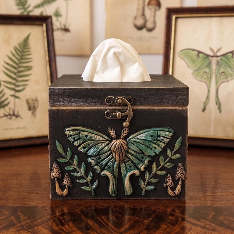 Puede incluir: Una caja de pa&ntilde;uelos cuadrada de madera negra con un dise&ntilde;o en relieve de una polilla luna verde, ramas con hojas y peque&ntilde;os hongos en el panel frontal. La caja incluye un cierre met&aacute;lico en tono bronce y un pa&ntilde;uelo de papel blanco.