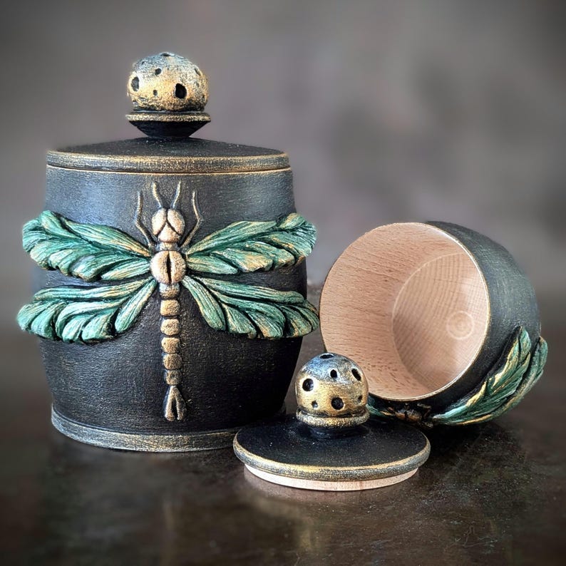 May include: A black cylindrical container with a textured finish and a gold-toned dragonfly relief on the front. It includes a matching lid with a small, perforated knob. A second, open container with a natural wood interior is placed beside it.