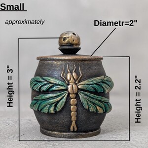 May include: A small dark jar with a lid featuring a gold and green dragonfly relief. The jar has a height of 3 inches, a lid diameter of 2 inches, and a body height of 2.2 inches. The surface has a textured, metallic finish with gold accents.