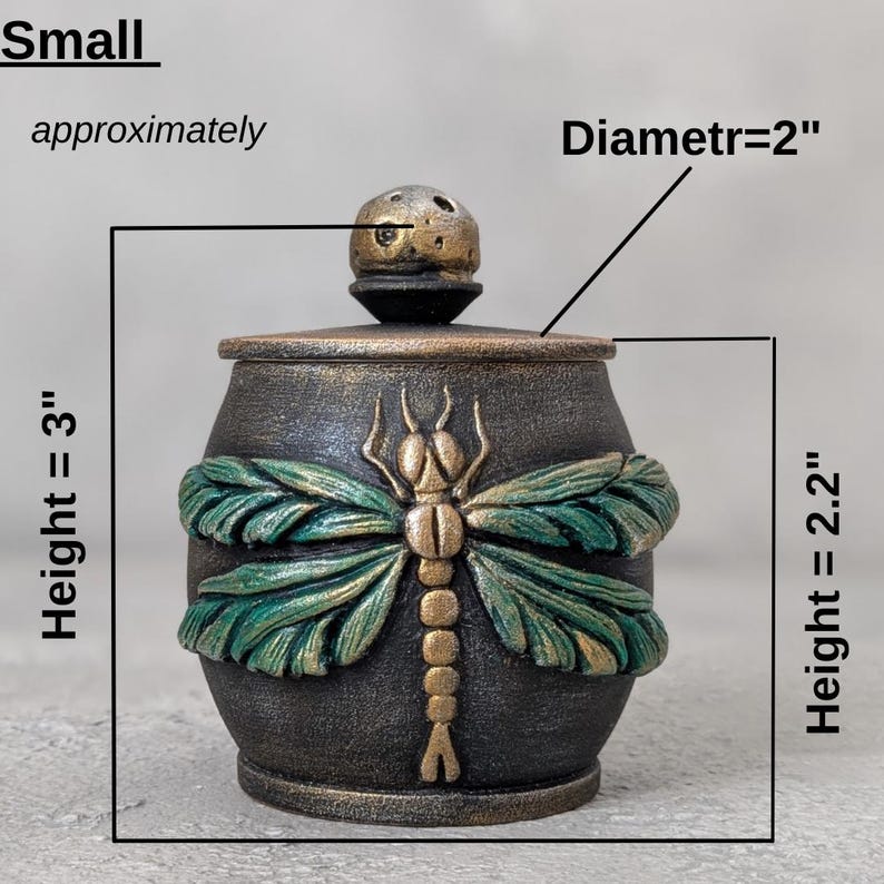 May include: A small dark jar with a lid featuring a gold and green dragonfly relief. The jar has a height of 3 inches, a lid diameter of 2 inches, and a body height of 2.2 inches. The surface has a textured, metallic finish with gold accents.