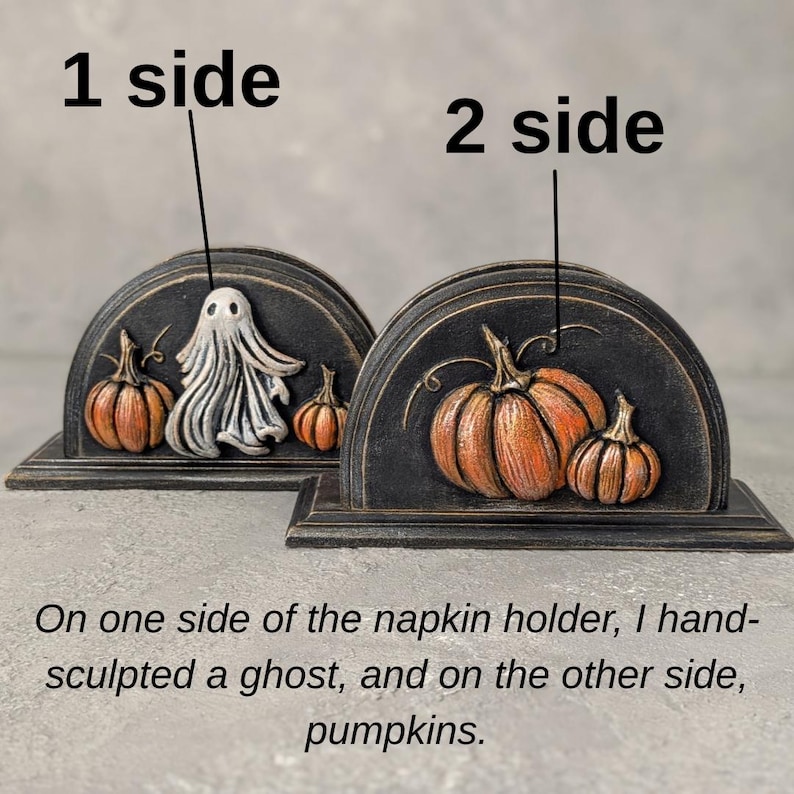 May include: A black semi-circular napkin holder featuring two distinct sides. One side displays a sculpted white ghost flanked by two orange pumpkins. The other side features two orange pumpkins. Text labels 1 side and 2 side are visible.