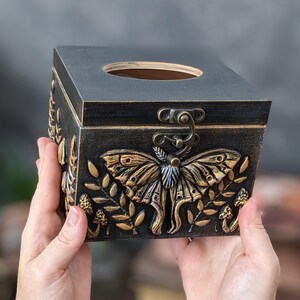 Puede incluir: Una caja cuadrada de madera negra con tapa abatible y una abertura circular en la parte superior. El frente presenta un relieve dorado de una gran polilla rodeada de elementos bot&aacute;nicos y peque&ntilde;os hongos. Un pestillo de metal color bronce asegura la tapa.
