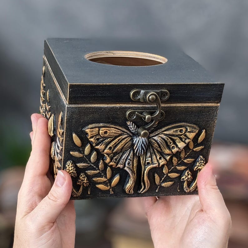 Puede incluir: Una caja cuadrada de madera negra con tapa abatible y una abertura circular en la parte superior. El frente presenta un relieve dorado de una gran polilla rodeada de elementos bot&aacute;nicos y peque&ntilde;os hongos. Un pestillo de metal color bronce asegura la tapa.
