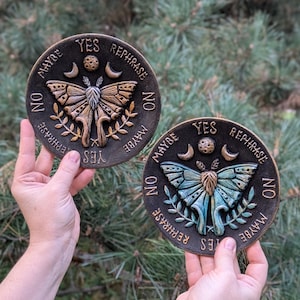 May include: Two circular plaques featuring a central moth design with raised wings, flanked by crescent moons and a small sphere. The text YES, NO, MAYBE, and REPHRASE is embossed around the perimeter. One plaque is bronze-toned, the other is teal.