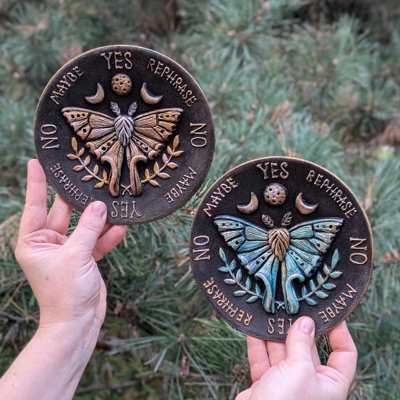 May include: Two circular plaques featuring a central moth design with raised wings, flanked by crescent moons and a small sphere. The text YES, NO, MAYBE, and REPHRASE is embossed around the perimeter. One plaque is bronze-toned, the other is teal.
