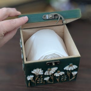 May include: A small, open wooden box with a dark green exterior and a hinged lid. The box is decorated with white floral relief carvings. Inside, a roll of white paper is visible. The box is being opened by a hand.