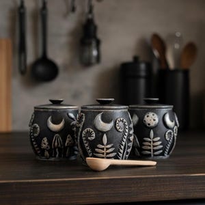 Peut inclure: Trois pots en céramique noire avec couvercles, chacun présentant des motifs en relief de couleur claire représentant des croissants de lune, des pleines lunes, des fougères et des champignons. Une petite cuillère en bois clair est posée.