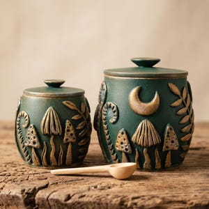 May include: Two dark green ceramic canisters with lids, featuring raised gold-toned relief designs of mushrooms, ferns, and a crescent moon. A small, light-colored wooden scoop rests in front of the canisters on a textured wooden surface.