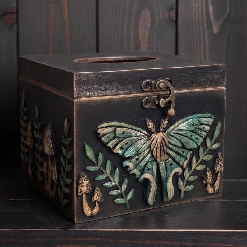 Puede incluir: Una caja de madera negra para pa&ntilde;uelos con un relieve de una polilla verde con detalles dorados en el panel frontal. El dise&ntilde;o incluye setas y ramas con hojas esculpidas en los laterales, junto con un cierre met&aacute;lico en la tapa frontal.