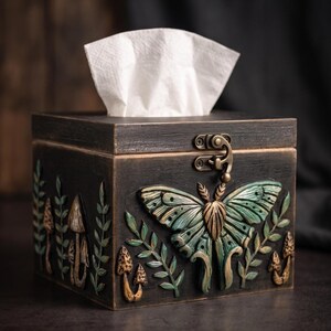 Puede incluir: Una caja de madera cuadrada para pa&ntilde;uelos con un acabado oscuro y desgastado. El frente presenta una polilla esculpida en relieve de color verde y bronce bajo un pestillo met&aacute;lico, flanqueada por tallas de peque&ntilde;os hongos y ramas.