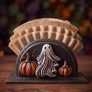 May include: A semi-circular black holder containing a stack of light brown paper coffee filters. The front of the holder features a raised, sculpted design of a central white ghost flanked by two orange pumpkins on a dark, textured background.