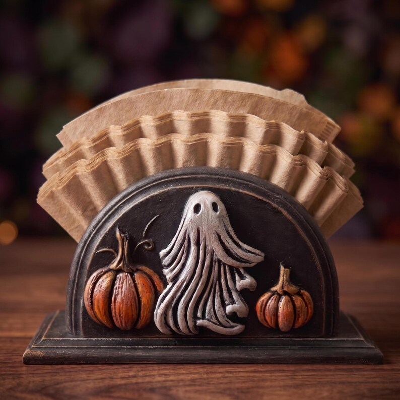 May include: A semi-circular black holder containing a stack of light brown paper coffee filters. The front of the holder features a raised, sculpted design of a central white ghost flanked by two orange pumpkins on a dark, textured background.