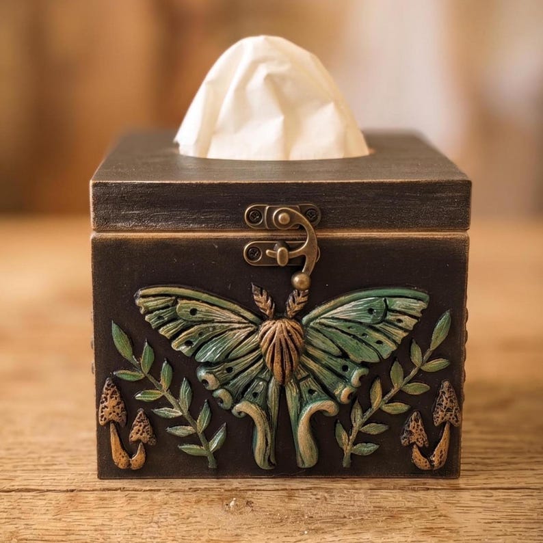 Puede incluir: Una caja de pa&ntilde;uelos cuadrada de madera marr&oacute;n oscuro con un relieve central de una polilla verde con las alas extendidas. El dise&ntilde;o incluye ramas con hojas esculpidas y peque&ntilde;os hongos a ambos lados de la figura central.