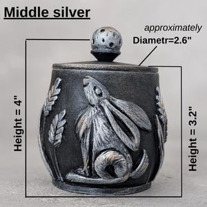 Puede incluir: Un tarro decorativo negro y plateado con tapa, con un relieve detallado de un conejo y espigas de trigo. El tarro mide aproximadamente 10 cm de alto, con un di&aacute;metro de 6,6 cm. La altura de la base es de 8,1 cm. El texto "Middle silver" est&aacute; en la parte superior.