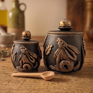 Puede incluir: Dos tarros de cer&aacute;mica gris oscuro con dise&ntilde;os en relieve de conejos en color bronce y tapas a juego. Una peque&ntilde;a cuchara de madera descansa frente a los tarros. Los tarros son probablemente para almacenar especias u otros art&iacute;culos de cocina.