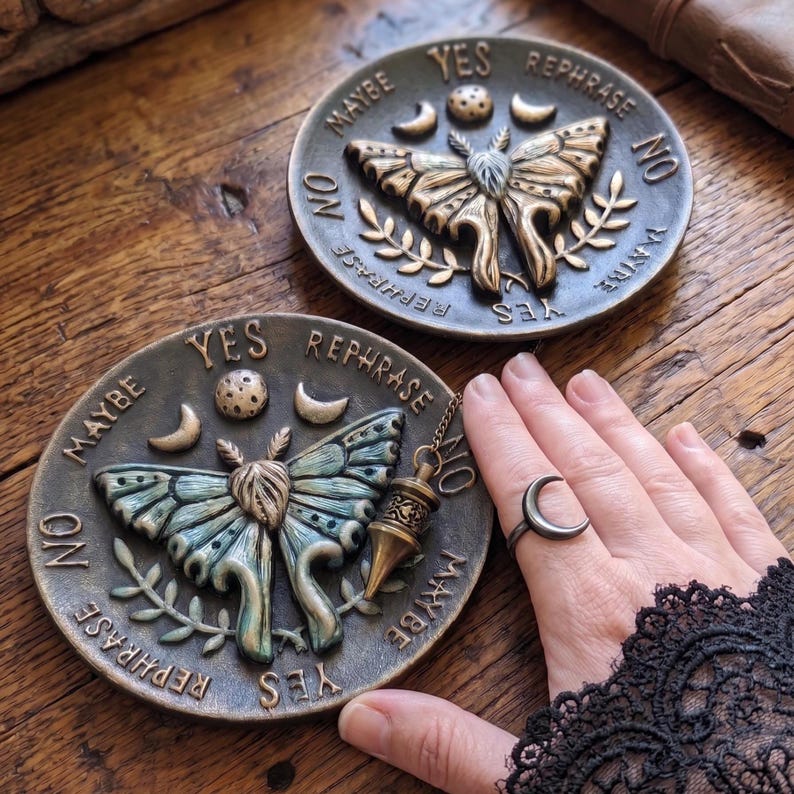 May include: Two circular bronze-toned divination boards featuring a central moth relief, celestial symbols including a moon and stars, and the words YES, NO, MAYBE, and REPHRASE. A metal pendulum hangs over one board, and a crescent moon ring is worn.