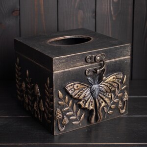 Puede incluir: Una caja para pa&ntilde;uelos cuadrada con acabado en bronce oscuro, que presenta un emblema de polilla en relieve en el frente, rodeado de motivos de setas y helechos. La tapa incluye una abertura ovalada central y un cierre met&aacute;lico frontal.