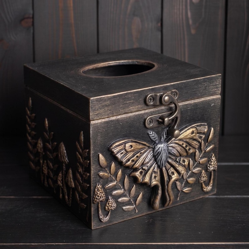 Puede incluir: Una caja para pa&ntilde;uelos cuadrada con acabado en bronce oscuro, que presenta un emblema de polilla en relieve en el frente, rodeado de motivos de setas y helechos. La tapa incluye una abertura ovalada central y un cierre met&aacute;lico frontal.
