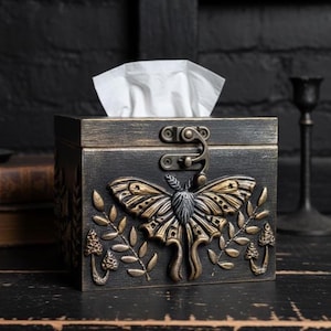 Puede incluir: Una caja de pa&ntilde;uelos rectangular de madera negra con un acabado dorado desgastado. El frente presenta un relieve de una polilla grande centrada entre dos ramas de hojas y peque&ntilde;os hongos. Un pestillo de metal asegura la tapa superior.