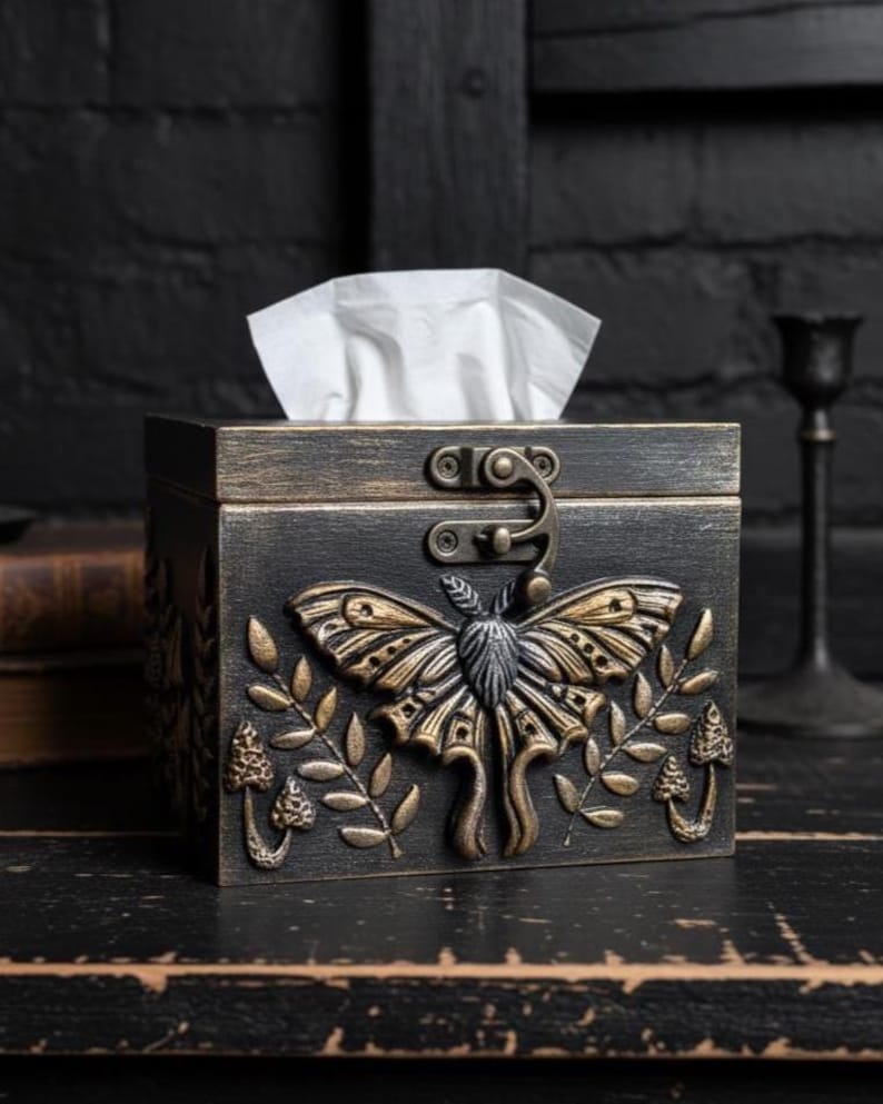 Puede incluir: Una caja de pa&ntilde;uelos rectangular de madera negra con un acabado dorado desgastado. El frente presenta un relieve de una polilla grande centrada entre dos ramas de hojas y peque&ntilde;os hongos. Un pestillo de metal asegura la tapa superior.